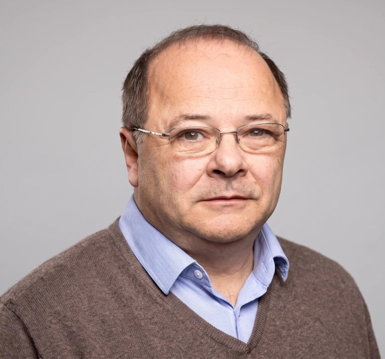A professional headshot of a middle-aged man with glasses, wearing a brown v-neck sweater over a light blue collared shirt, set against a solid gray background.