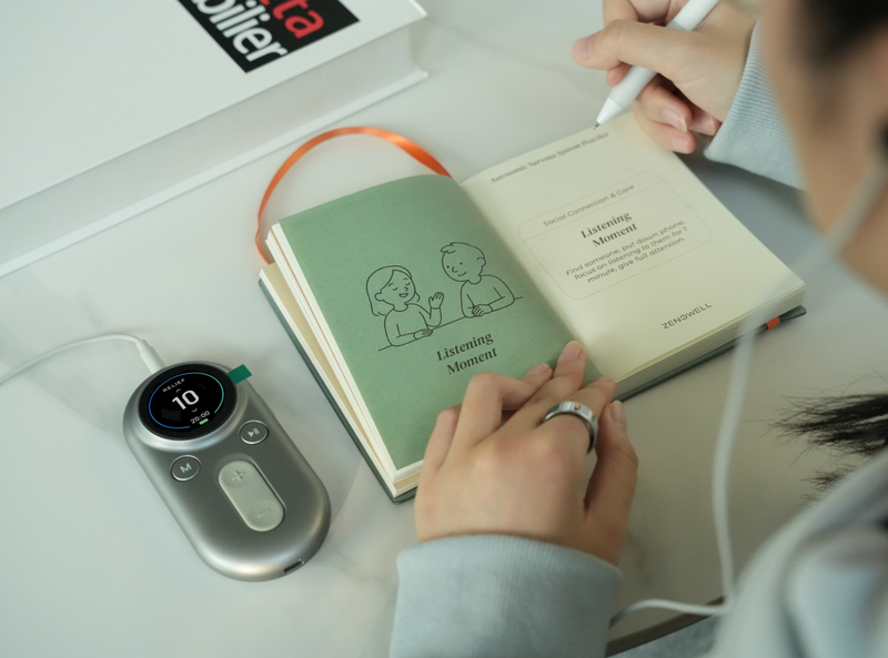 ZenoWell wearable device luna placed beside an open ‘Listening Moment’ practice journal, with a person writing notes at a desk.