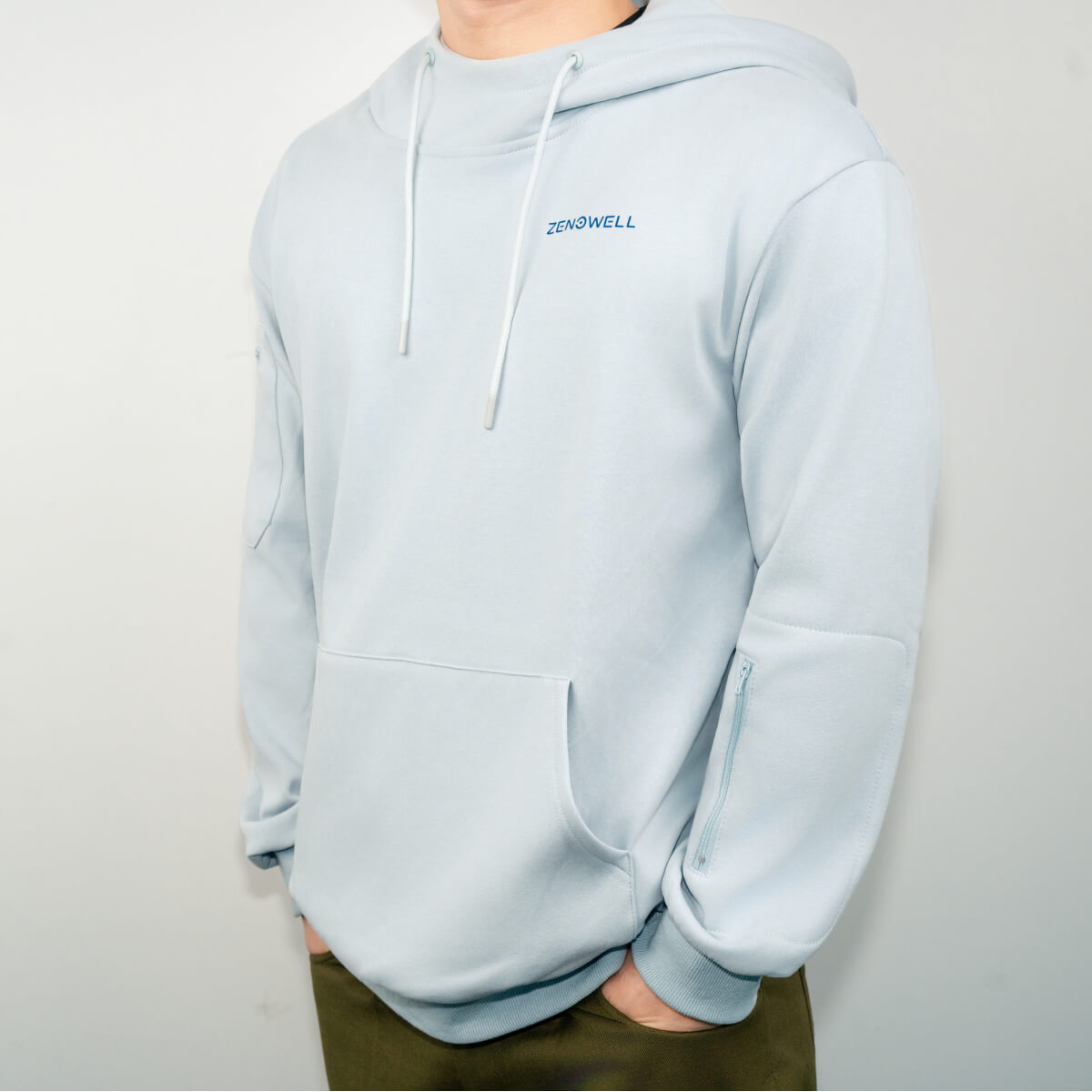 A person wearing a light blue ZenoWell hoodie with the brand logo on the chest and a zippered pocket on the sleeve.