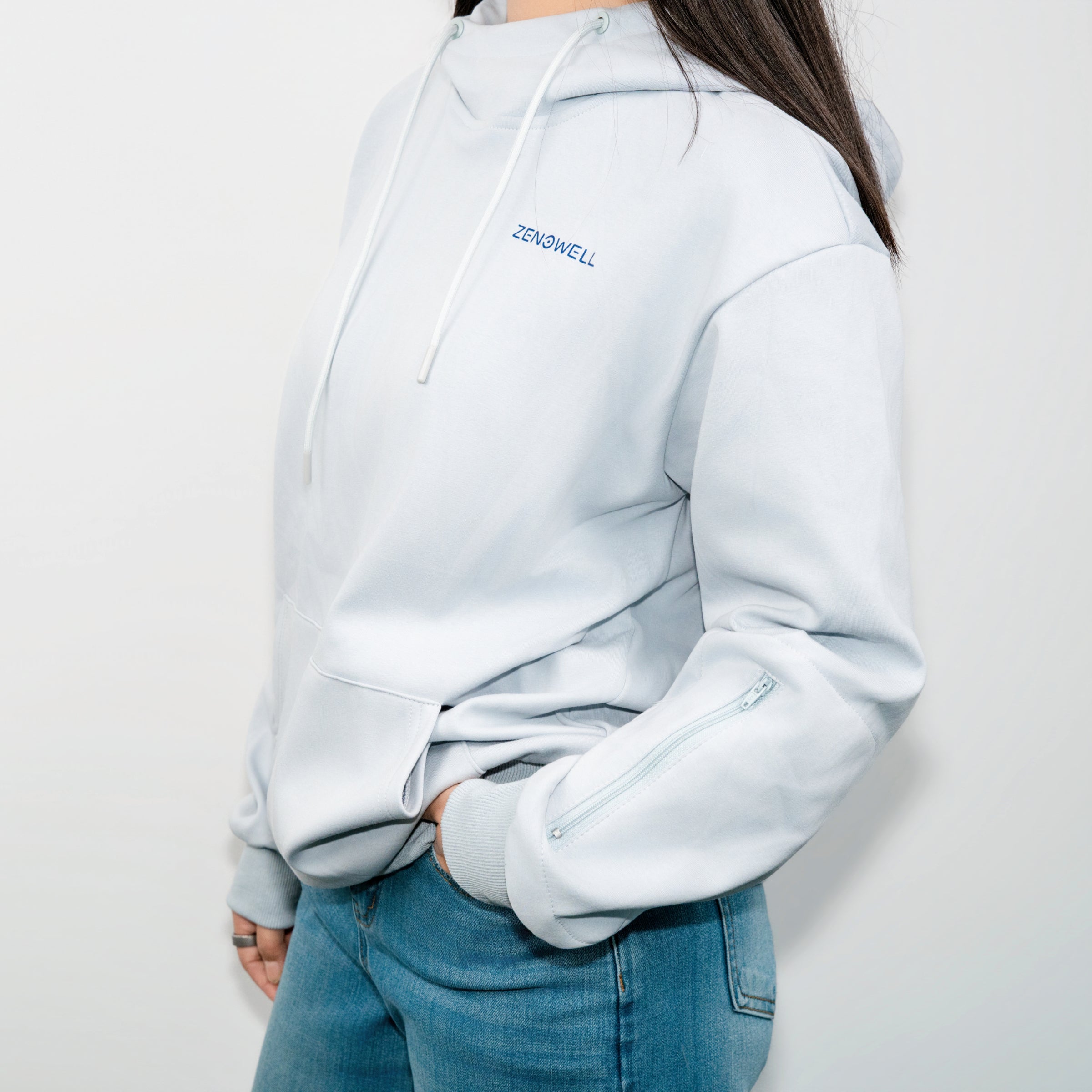 A person wearing a light blue ZenoWell hoodie, with one hand tucked into the pocket and showcasing the branding on the chest.